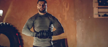 Muscular man in gray rashguard and black MMA gloves posing in dimly lit training gym with tire in background