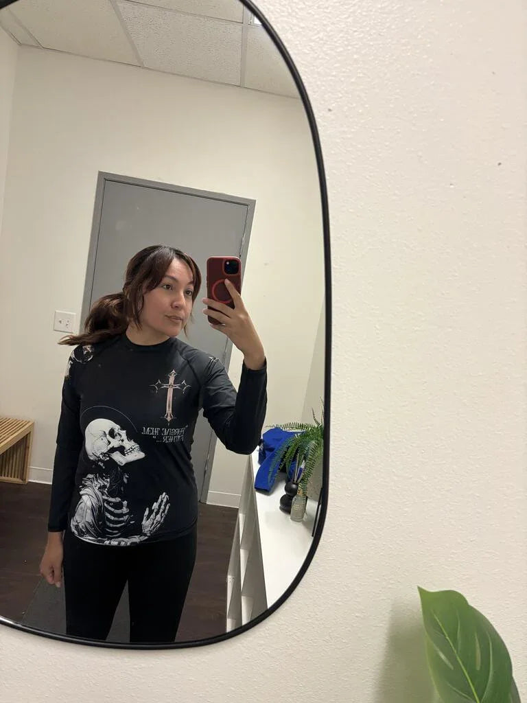 woman taking mirror selfie wearing black long sleeve shirt with praying skeleton and cross graphic in simple room